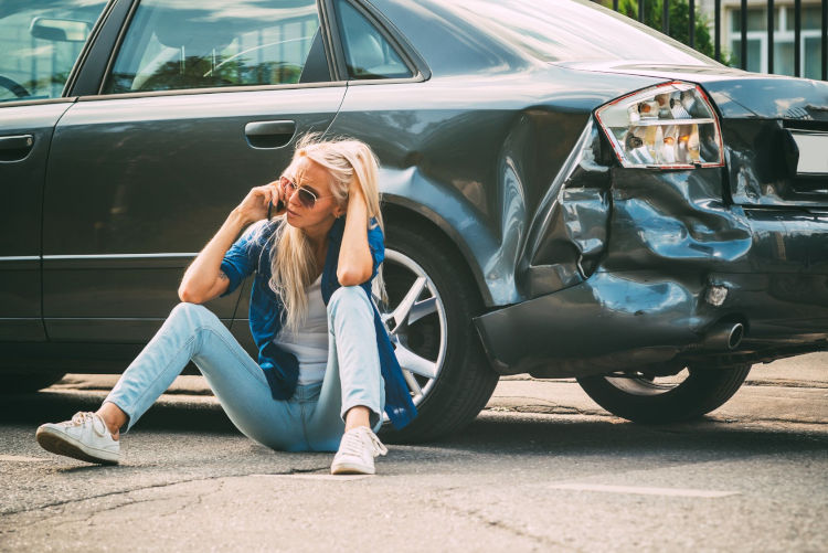 Verzweifelte Frau sitzt auf dem Boden vor ihrem beschädigten Fahrzeug. Sie ruft einen KFZ Gutachter in Göttingen an, weil sie unverschuldet ist und eine Dokumentation des Schadens brauch.